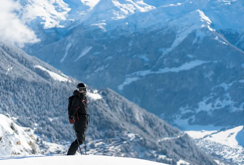 A skier looks towards the camera, standing still against a good looking backdrop of snowy trees on a facing slope across the valley